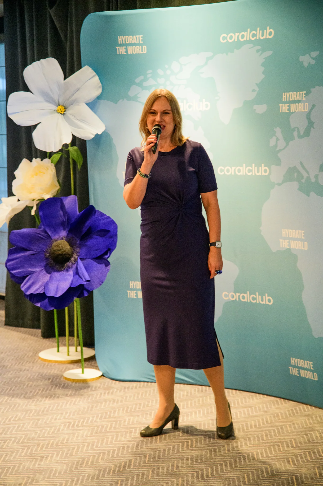 Gosia w granatowej sukience mówi do mikrofonu, za nią ścianka z mapą świata i duże papierowe kwiaty